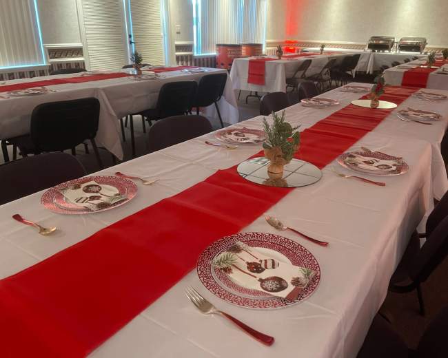 A dining setup features multiple tables covered with white tablecloths and red runners, arranged with decorative plates and utensils.