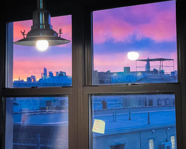 A pendant light hangs above a window, which frames a colorful sunset skyline and neighboring rooftops.