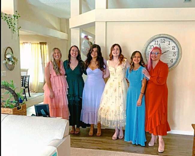 A group of six women stands together in a spacious, well-lit room with light-colored walls and a large wall clock.