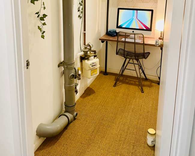 The image shows a small home office nook with a desk, chair, and computer, surrounded by pipes and greenery on the walls.