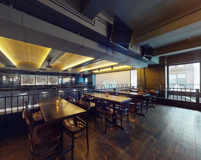 The image shows an interior view of a restaurant with wooden tables and chairs, dim lighting, and a large screen on one wall.