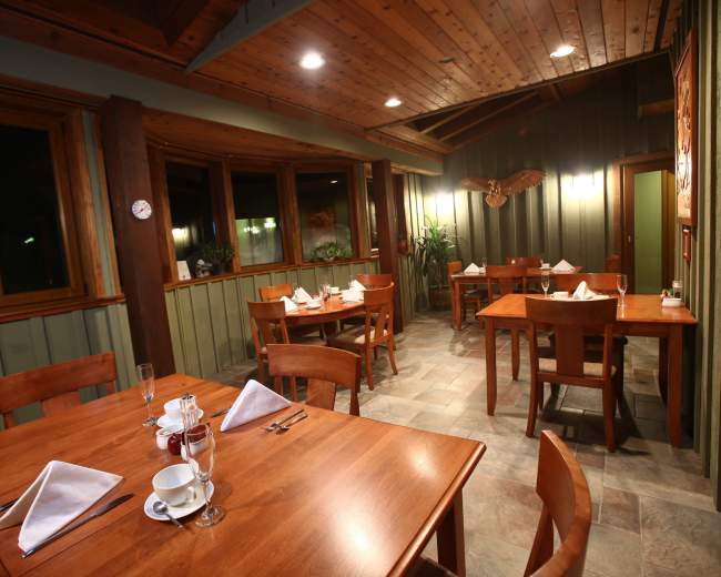 The image shows a dimly lit restaurant interior with wooden tables set for dining and large windows allowing for natural light.