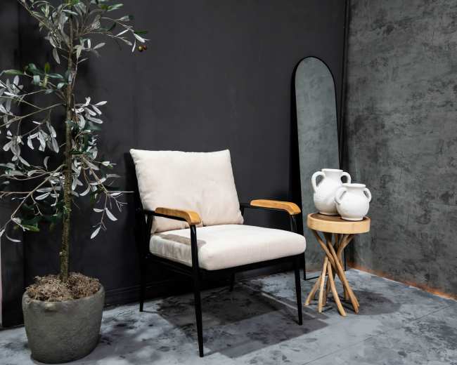 A modern interior features a plush armchair with wooden armrests beside a round wooden side table holding two ceramic pitchers, all against a dark gray wall.
