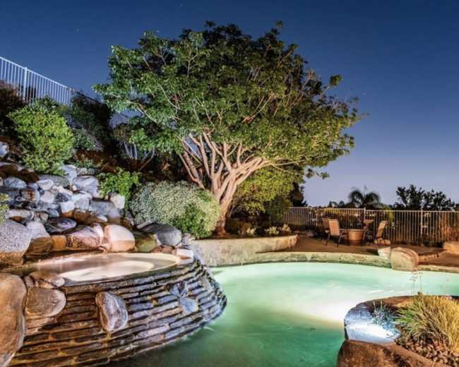 A illuminated backyard features a stone hot tub beside a pool and a large tree at dusk.