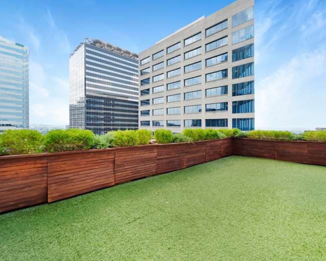 A rooftop terrace with artificial grass and wooden fencing, overlooking adjacent high-rise buildings.