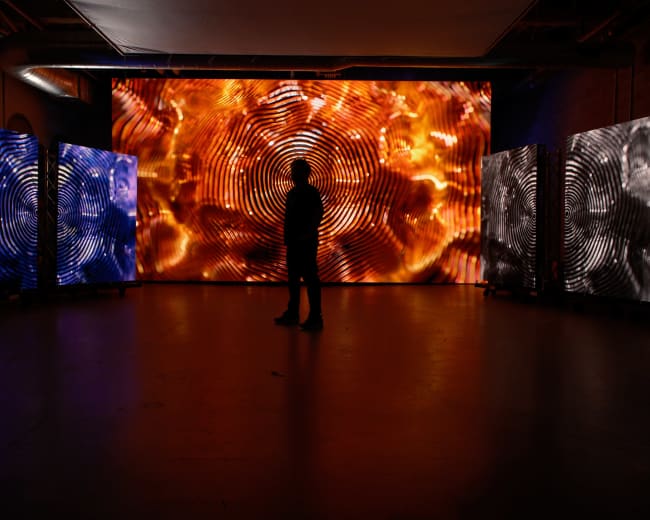 A person stands silhouetted in a dimly lit room, facing a large screen displaying vibrant, swirling patterns, flanked by two smaller screens with contrasting visuals.