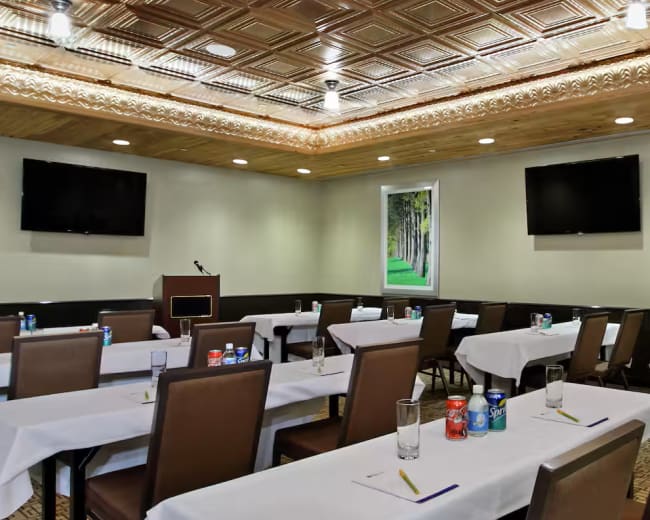 The image depicts a conference room set up with rows of tables covered in white tablecloths, accompanied by chairs, and two large television screens mounted on the walls.