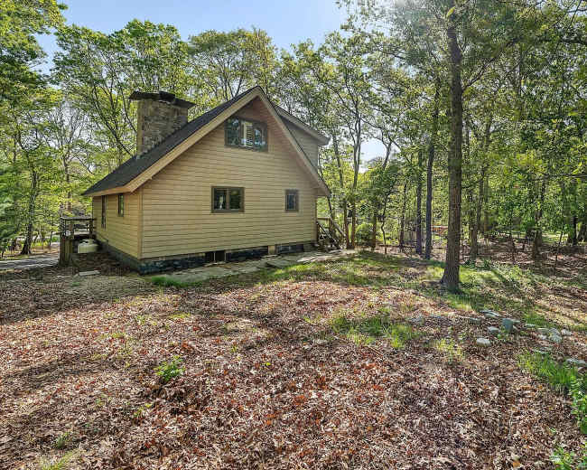 A wooden house with a steep roof is set among trees in a clearing, surrounded by fallen leaves.