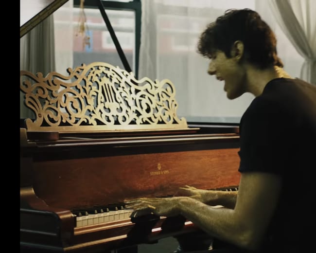 A young man is playing a wooden piano in a well-lit room with large windows.