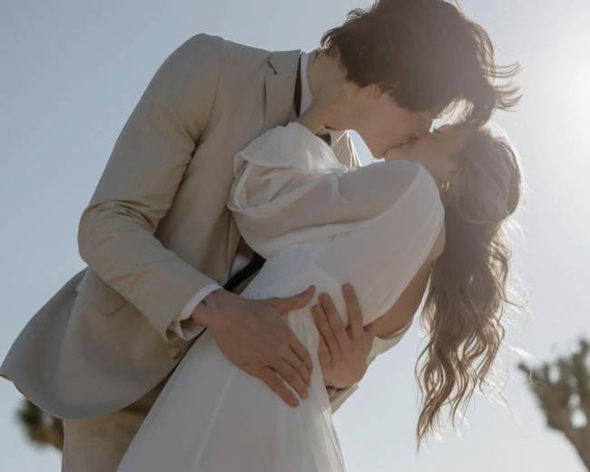 A couple kisses each other in a desert landscape while the sun shines overhead.