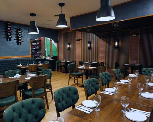 The interior of a restaurant features wooden tables arranged with plates and glasses, surrounded by green upholstered chairs and walls with a mix of textures.