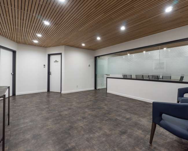 The image shows a modern office space with a wooden ceiling, a glass-walled conference room, and a seating area featuring a blue armchair.