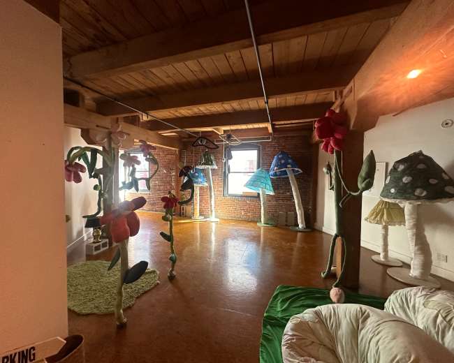 The room features large, colorful fabric sculptures resembling flowers and mushrooms, set against a backdrop of exposed brick walls and wooden beams.