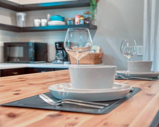 A wooden dining table is set with two place settings, each featuring a white bowl, a plate, a fork, and a glass, with a kitchen backdrop visible.