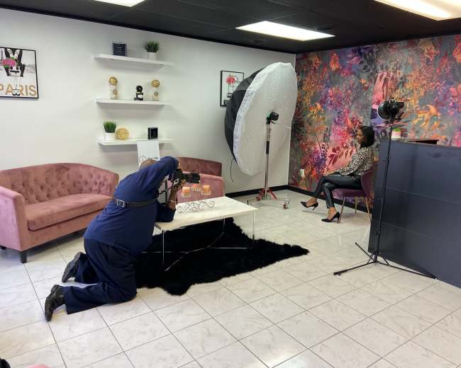 A photographer is kneeling on the floor while capturing a portrait of a seated model in a studio decorated with flowers and modern furnishings.