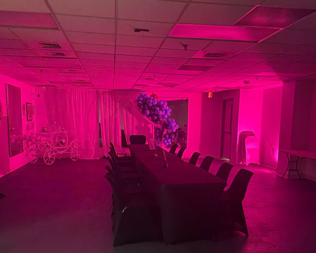 A large table is set up in a dimly lit room with pink lighting and a decorative balloon arrangement in the background.
