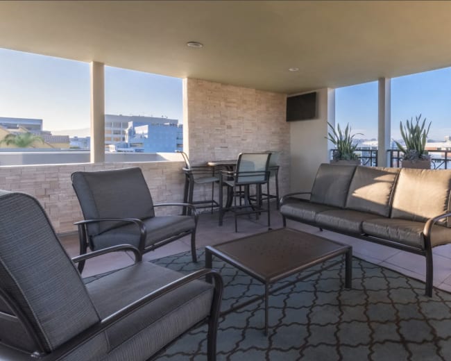 The image shows a modern balcony area with several metal-framed chairs and a table, surrounded by large windows and decorative plants.