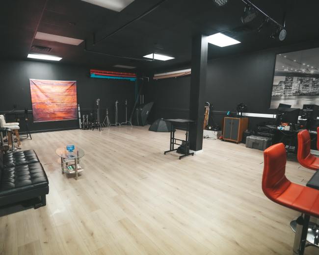 The image shows a spacious, dimly lit studio with wooden flooring, black walls, and various equipment laid out, including furniture and photography gear.