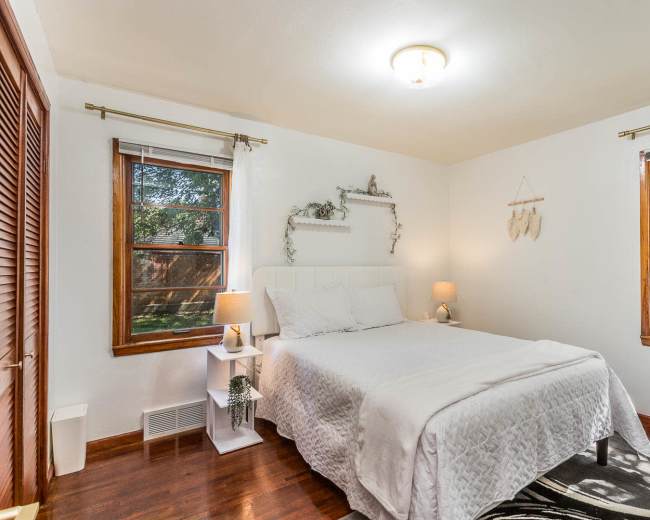 The room features a queen-sized bed with white bedding, two windows, and a small nightstand with lamps.
