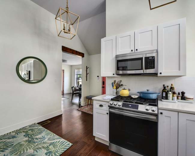 A modern kitchen with white cabinets, stainless steel appliances, and a round mirror on the wall, opens into a well-lit living space.