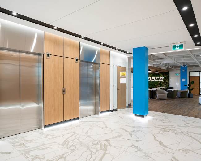 The image shows a modern lobby area with wooden elevator doors, a sleek metal elevator, and a blue wall featuring a sign, along with a marble floor and seating area in the background.