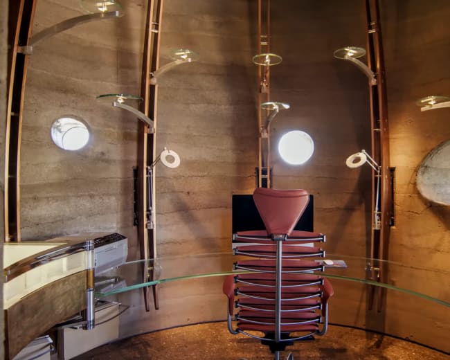 The image shows a modern, circular room featuring a glass table and a stack of metal chairs, illuminated by several wall-mounted lights and having two round windows.