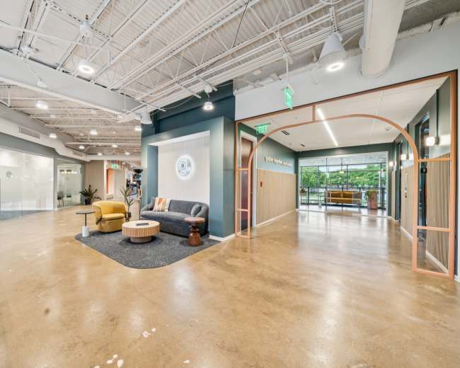 The image shows a modern office hallway with a seating area featuring a gray sofa, yellow chair, and potted plants, alongside glass-walled meeting rooms.