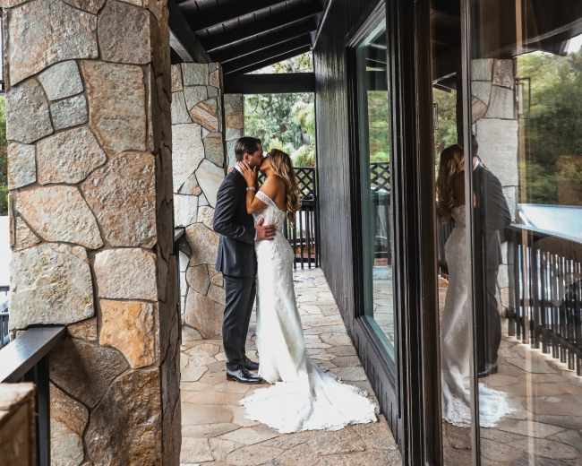 A couple kisses on a stone balcony under a wooden overhang, with reflections visible in large glass panels.