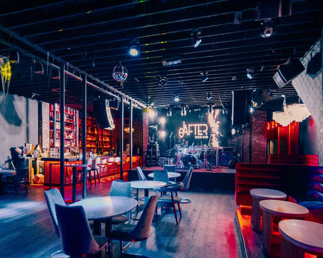 The image shows an empty nightclub interior featuring a bar area, tables, and a stage with lighting elements.