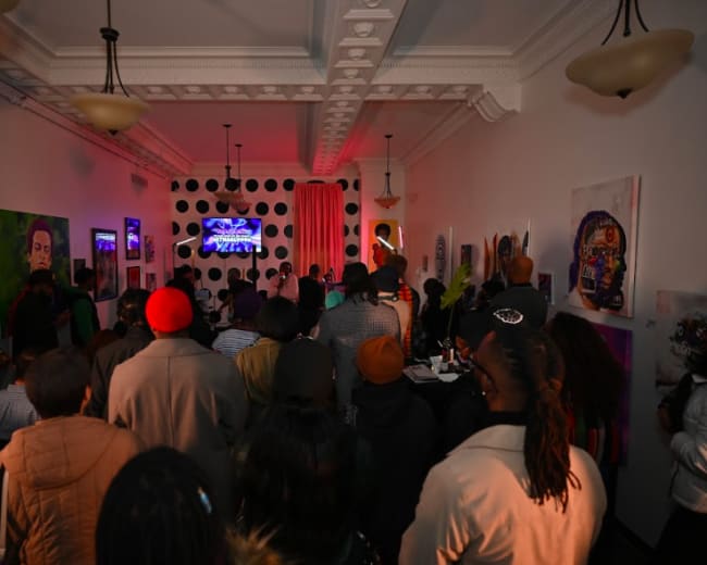 A crowd of people gathers in a dimly lit room adorned with colorful artwork, facing a screen for a performance or presentation.