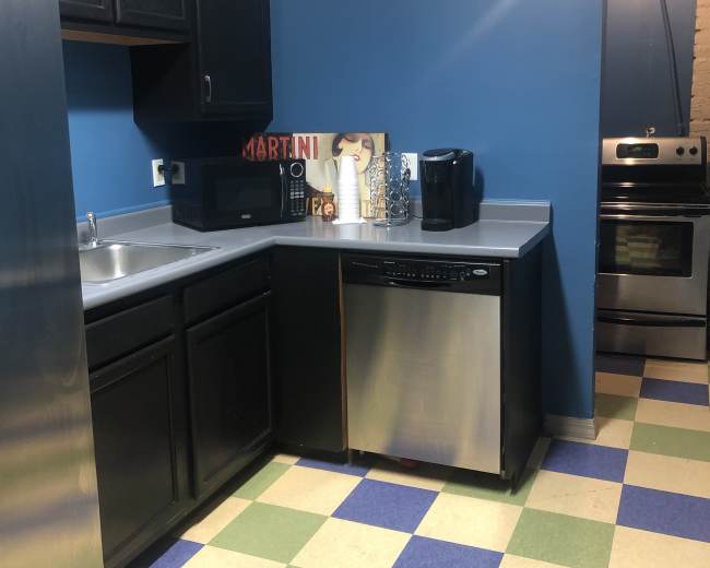 The image shows a kitchen with dark cabinets, a stainless steel dishwasher, a microwave, and a coffee maker on a countertop, set against a blue wall and checkered green and blue floor tiles.