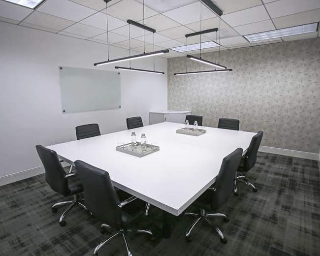 A modern conference room features a large white table surrounded by black chairs, with minimalistic light fixtures above and a patterned wall in the background.