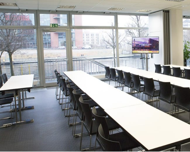 The image shows a modern conference room with rows of tables and chairs set up for a meeting, and large windows overlooking a canal.