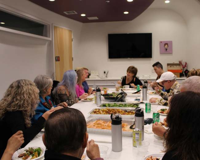 A group of people is seated around a long table, enjoying a meal together in a brightly lit meeting room.