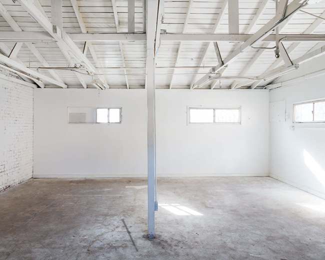 The image shows an empty, well-lit room with white walls, a concrete floor, and two small windows.