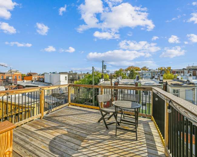 A wooden deck with two chairs overlooks a cityscape under a blue sky with scattered clouds.