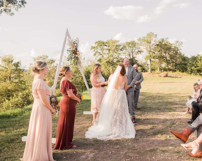 A couple exchanges vows during an outdoor wedding ceremony, surrounded by guests seated on either side and a decorated arch in the background.