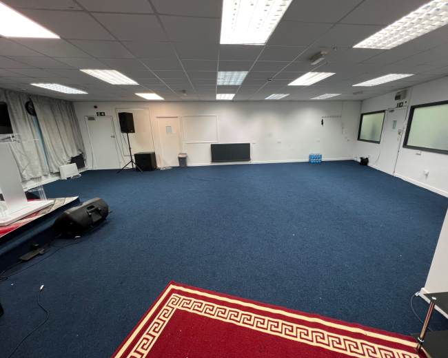 The image shows a spacious, empty room with blue carpet, a red rug at the front, and plain walls.