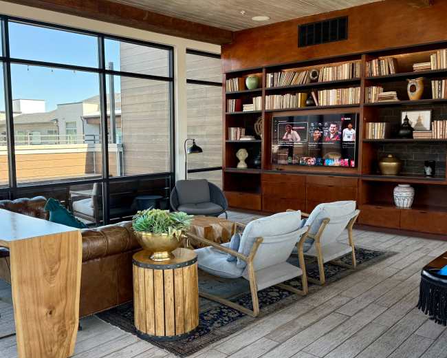 The image shows a modern lounge area featuring a large leather sofa, armchairs, a wooden dining table, bookshelves, and a view of an outdoor space through large windows.