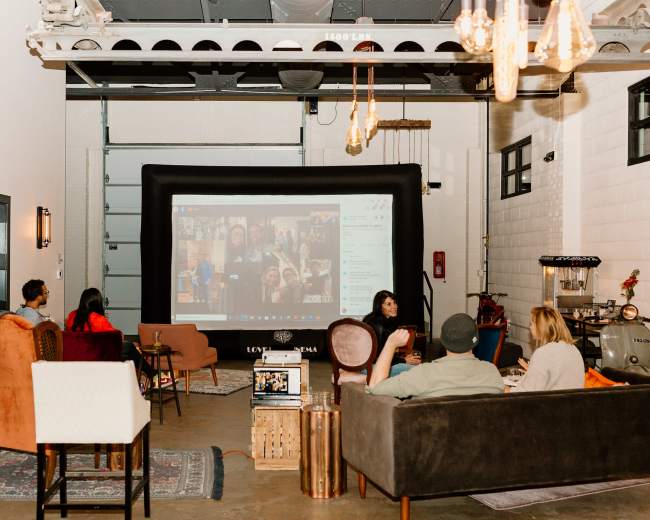 A group of people is seated in a warehouse-style space with a large screen displaying a video call.