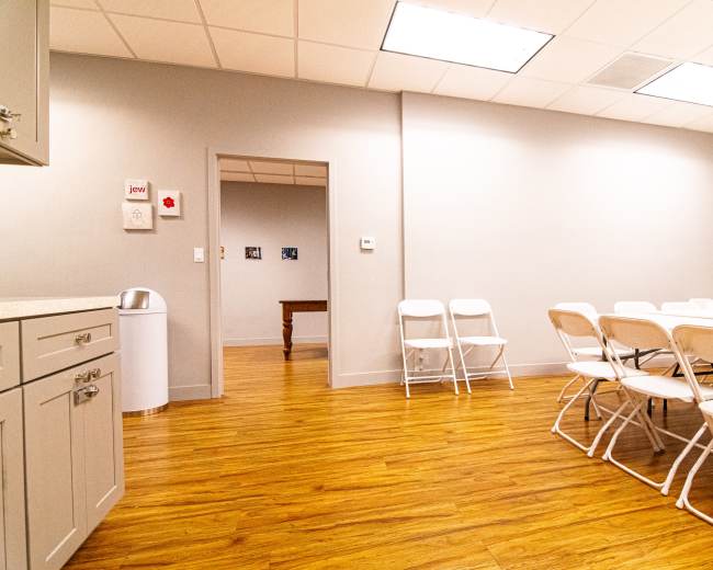 A brightly lit meeting room features a long table with white folding chairs arranged around it, and a doorway leading to another room in the background.