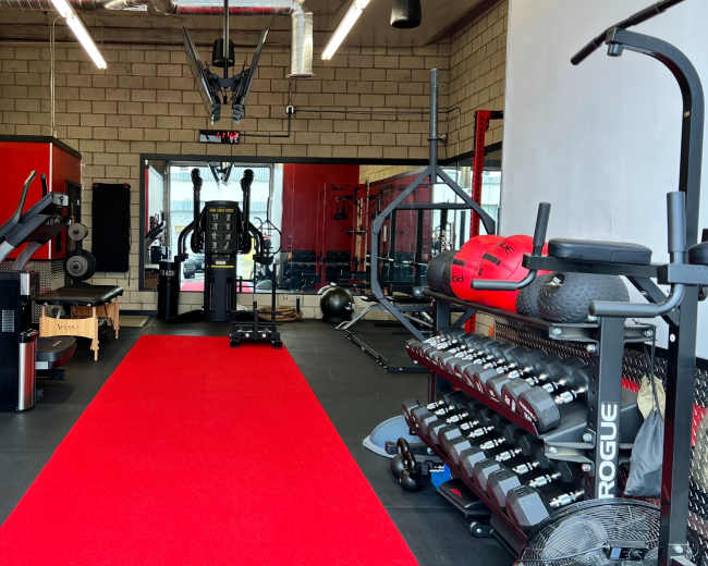 The image shows a modern gym interior with exercise equipment, including weights, a cable machine, and a red carpet leading to a mirrored wall.