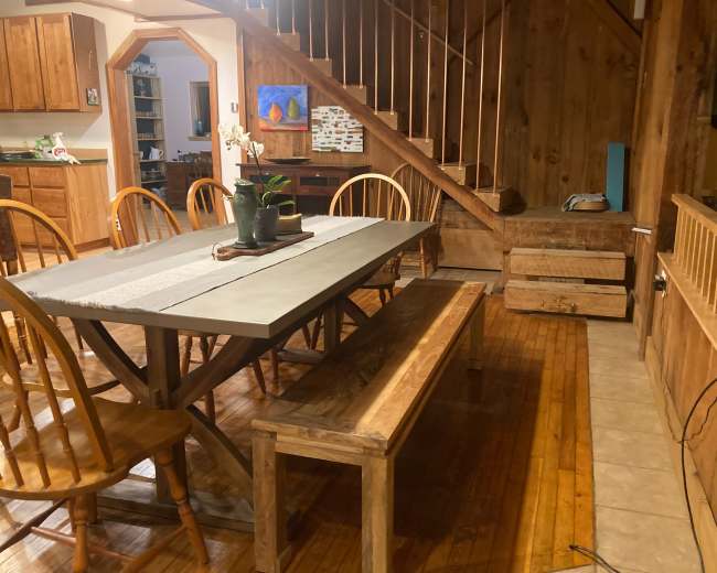 A wooden dining area with a large rectangular table and benches, situated next to a staircase and a wall featuring wooden paneling.