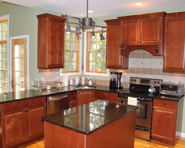 The image shows a modern kitchen with wooden cabinets, black granite countertops, and stainless steel appliances, set in a light-filled environment with large windows.