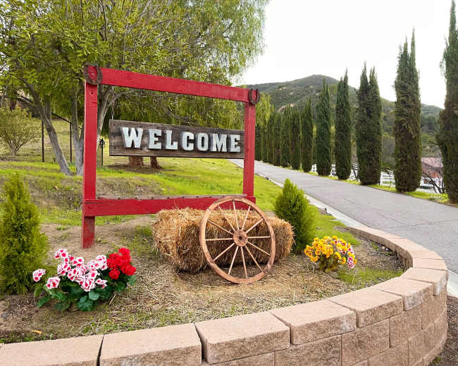 A red wooden welcome sign stands at the entrance of a landscaped area, surrounded by flowers and a wagon wheel along a paved road lined with tall cypress trees.