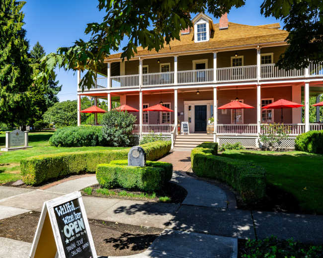 The image shows a two-story pink building with white railings and red umbrellas on the porch, surrounded by manicured lawns and trees.