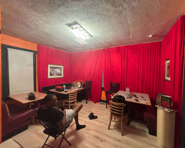 The image shows a small room with red curtains and walls, featuring two tables, chairs, and scattered personal items.