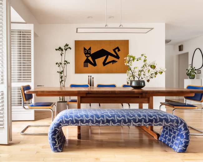 A modern dining area featuring a large wooden table, a blue and white patterned bench, and a wall adorned with a decorative artwork.