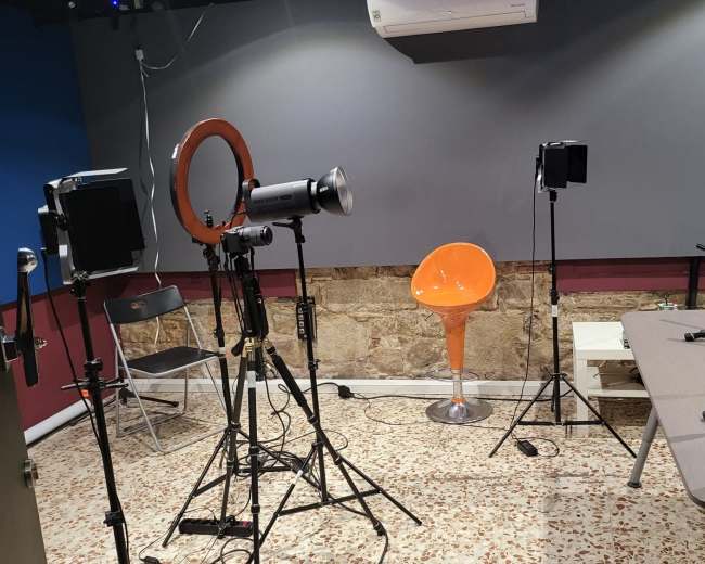 The image shows a studio setup with lighting equipment, an orange chair, a table with microphones, and a textured wall in the background.