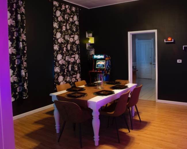 A dining room with a white table set for six, surrounded by wooden chairs, against a wall decorated with floral wallpaper and an arcade game machine in the background.